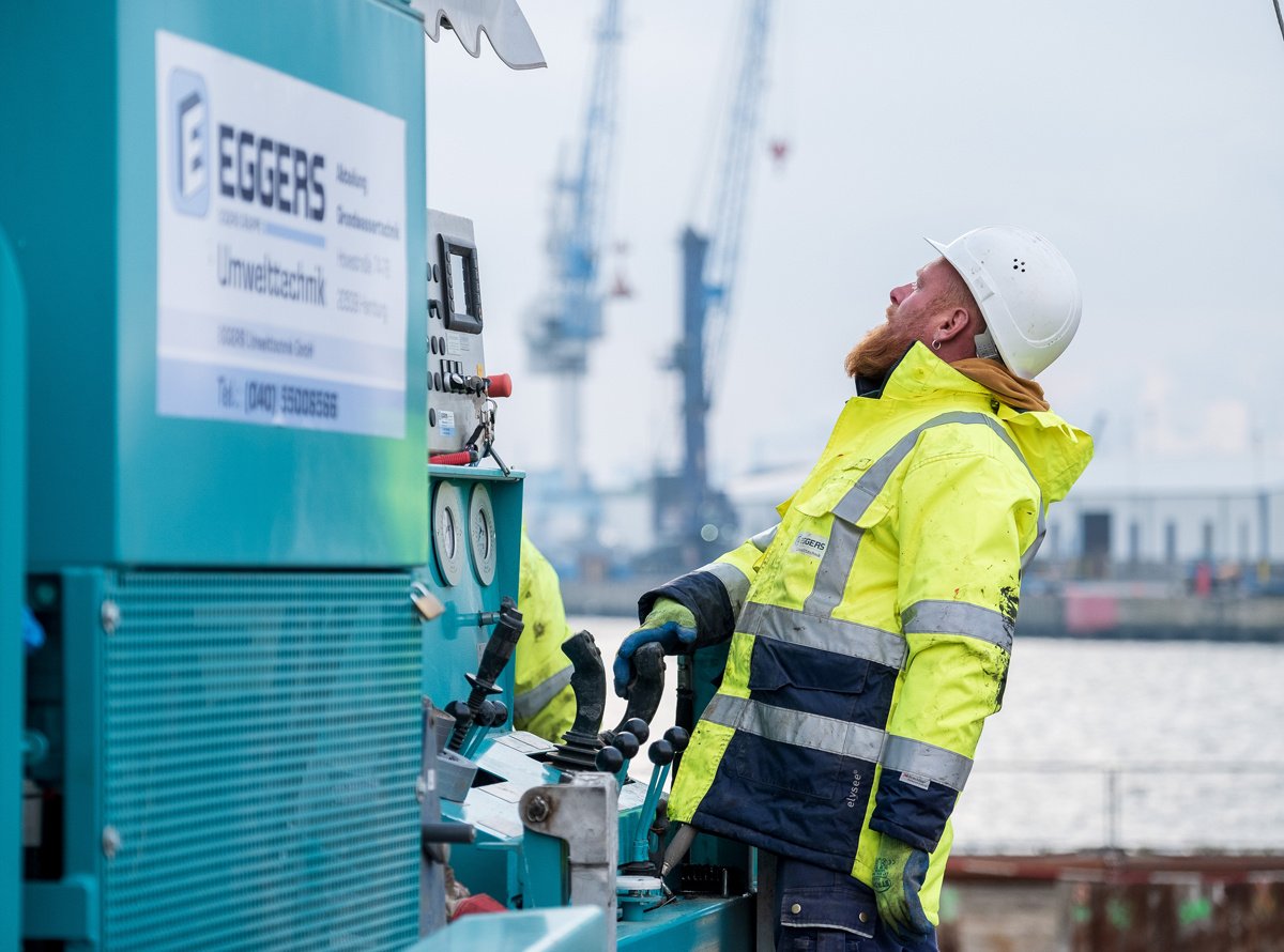 Ein Arbeiter der Firma EGGERS Umwelttechnik ist bei der Wartung oder Inspektion einer technischen Anlage zu sehen. Er trägt Sicherheitsausrüstung bestehend aus einem weißen Schutzhelm und einer gelben Warnschutzjacke mit reflektierenden Streifen. Der Mann blickt nach oben, während er an einer türkisfarbenen technischen Anlage arbeitet, Die das EGGERS-Logo trägt. Im unscharfen Hintergrund sind Hafenkräne zu erkennen, was darauf hindeutet, dass sich die Szene in einem Hafengebiet befindet. Das Bild vermittelt einen professionellen Eindruck von industrieller Wartungsarbeit unter Einhaltung von Sicherheitsstandards.