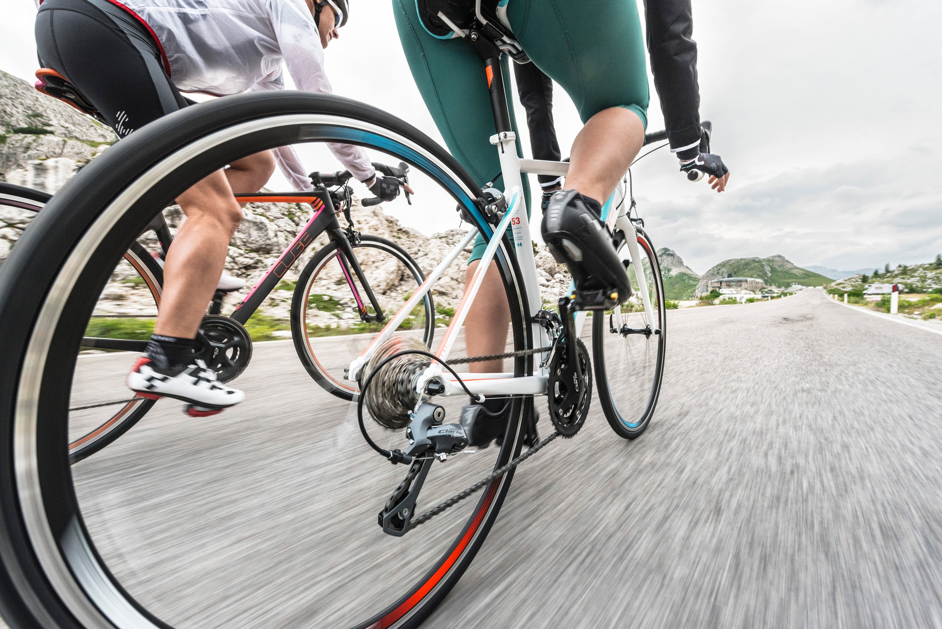 Dynamische Nahaufnahme von Rennradfahrern in Bewegung, aufgenommen aus einer niedrigen Perspektive mit Fokus auf die Räder und Beine. Ein Fahrer trägt eine grüne Radhose und schwarze Radschuhe, ein anderer ist in Weiß gekleidet. Die Fahrer bewegen sich auf einer Straße mit Bergen im Hintergrund. Das Bild vermittelt durch Bewegungsunschärfe am Boden Geschwindigkeit und zeigt technische Details wie Kettenblätter, Schaltung und Reifen mit roten und türkisfarbenen Akzenten. Die Aufnahme wurde bei bewölktem Himmel gemacht.