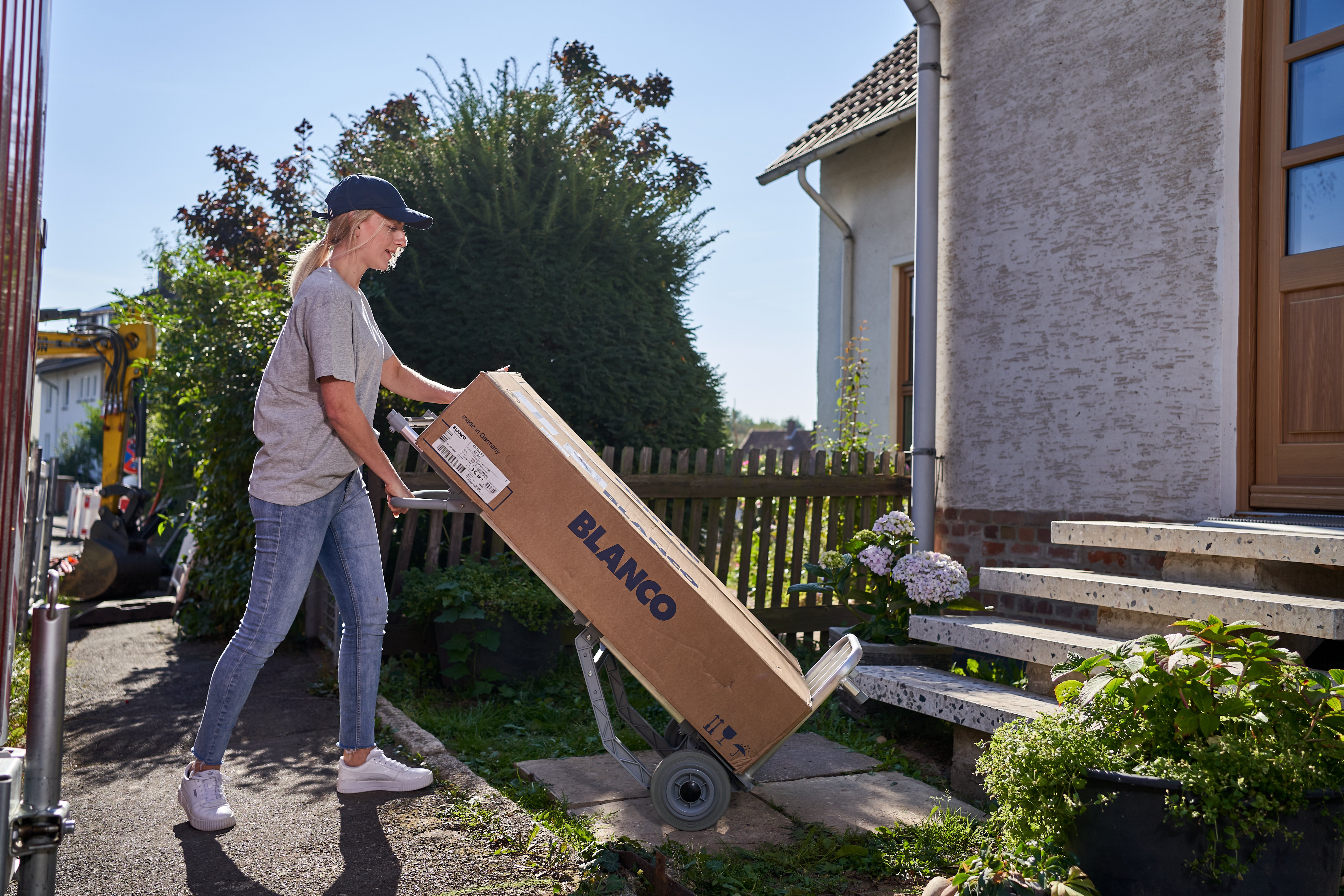 Eine Lieferantin in Baseballkappe, grauem T-Shirt und Jeans transportierte einen großen BLANCO-Karton auf einer Sackkarre die Stufen zu einem Hauseingang hinauf. Das Haus hat eine weiße Fassade und ist von einem Garten mit Hortensien und anderen Pflanzen umgeben.