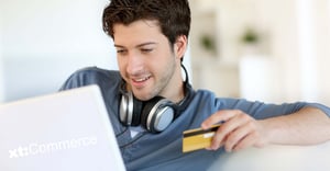 A smiling young man sits in front of a laptop. He is holding a credit card in his hand. The image implies an online transaction and symbolizes xt:Commerce.